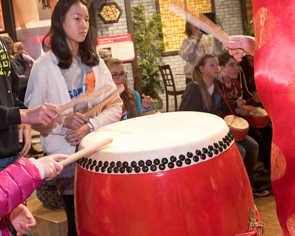 Children using drumsticks to play large drum in the Take Me There: China exhibit.